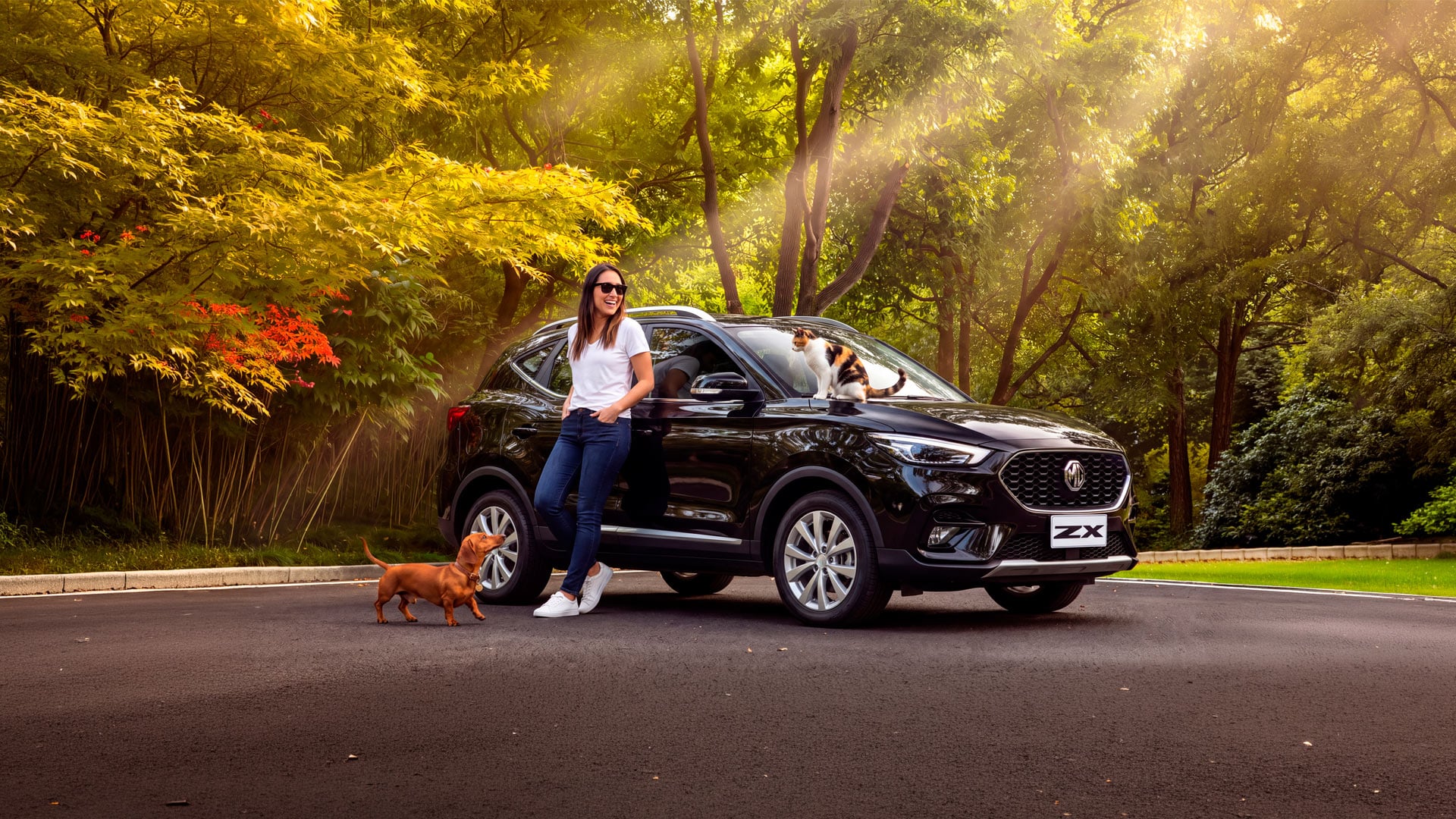 Mujer junto a un MG ZX negro con un perro salchicha y un gato sobre el capó, en un parque